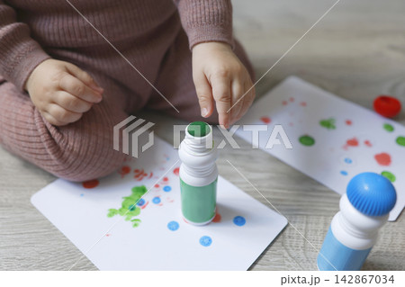 Toddler hands close-up touching green dot marker tip on drawing paper. Creative educational activity for fine motor skills development at home. Real indoor lifestyle cropped macro view 142867034