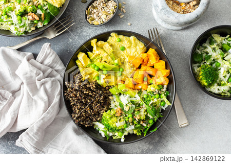 A quinoa bowl is arranged with roasted pumpkin, hummus, and a fresh salad. Plates are set on a table with utensils. Sunshine comes through the window A quinoa bowl is arranged with roasted pumpkin, hummus, and a fresh salad. Plates are set on a table with utensils. Sunshine comes through the window 142869122