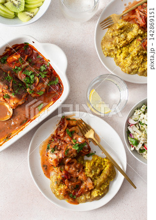 Plates hold amaranth, millet, and quinoa porridge served with baked white hake in tomato sauce. Fresh salad is beside the porridge. A bowl of kiwi sits nearby for dessert Plates hold amaranth, millet, and quinoa porridge served with baked white hake in tomato sauce. Fresh salad is beside the porridge. A bowl of kiwi sits nearby for dessert 142869151