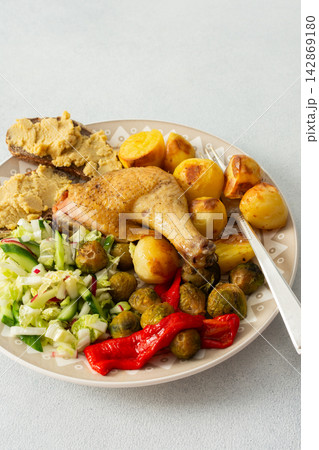 A plate holds baked chicken leg, potatoes, fresh salad, roasted Brussels sprouts, and chickpea bread. This meal is ready to eat at a dining table A plate holds baked chicken leg, potatoes, fresh salad, roasted Brussels sprouts, and chickpea bread. This meal is ready to eat at a dining table 142869180