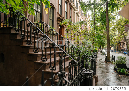 Side view of a historic New York brownstone stoop with ornate iron railings and stone steps along a wet sidewalk. A Ukrainian flag hangs above the entrance, adding cultural symbolism to the quiet tree 142870226