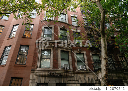 A classic red brick New York apartment building featuring decorative stone window frames and black metal fire escapes. Green trees frame the historic urban facade in a charming residential A classic red brick New York apartment building featuring decorative stone window frames and black metal fire escapes. Green trees frame the historic urban facade in a charming residential 142870241