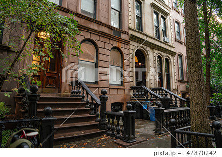 A row of classic brownstone townhouses with stone staircases and iron railings on a quiet New York City street. The residential buildings are framed by trees and urban greenery in a peaceful A row of classic brownstone townhouses with stone staircases and iron railings on a quiet New York City street. The residential buildings are framed by trees and urban greenery in a peaceful 142870242