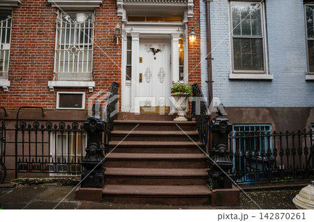 A traditional New York brownstone entrance featuring stone steps, ornate black iron railings, and a white decorative door. Wet sidewalk and brick facade highlight historic urban residential A traditional New York brownstone entrance featuring stone steps, ornate black iron railings, and a white decorative door. Wet sidewalk and brick facade highlight historic urban residential 142870261