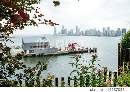 The Hudson River and Jersey City skyline are framed by green plants and pink flowers in the foreground. A waterfront pier and fireboat add urban detail to the soft, cloudy cityscape. The Hudson River and Jersey City skyline are framed by green plants and pink flowers in the foreground. A waterfront pier and fireboat add urban detail to the soft, cloudy cityscape. 142870284