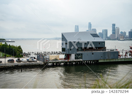 Modern waterfront building on a pier along the Hudson River with Manhattan skyline in the background. Urban riverfront and city skyscrapers create a recognizable New York City landscape. Modern waterfront building on a pier along the Hudson River with Manhattan skyline in the background. Urban riverfront and city skyscrapers create a recognizable New York City landscape. 142870294