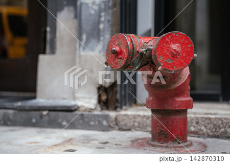 Detailed close-up of a weathered red fire hydrant on an urban sidewalk. Peeling paint and metal texture highlight city infrastructure and public safety equipment. 142870310