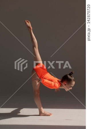 A little Caucasian girl gymnast in an orange leotard demonstrates her skills on a white background. A little Caucasian girl gymnast in an orange leotard demonstrates her skills on a white background. 142870720
