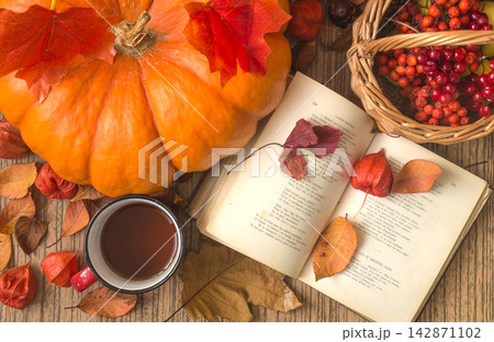 Autumn still life; open book, mug of tea; orange squash; basket with red berries and dry leaves on a wooden background top view 142871102
