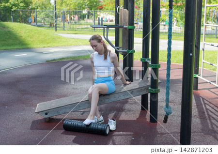 A Caucasian woman stretches her feet with a foam roller at a sports ground. A Caucasian woman stretches her feet with a foam roller at a sports ground. 142871203