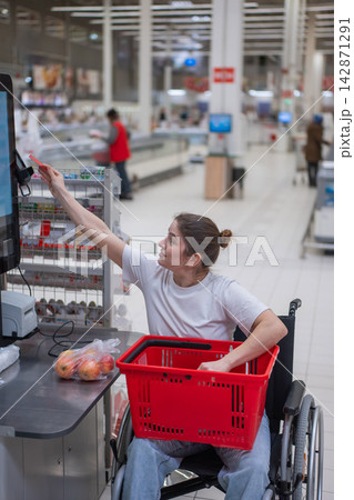 A Caucasian woman in a wheelchair pays for groceries at a self-checkout counter in a supermarket. 142871291