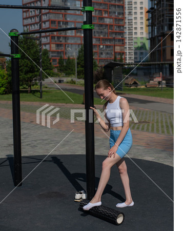 A Caucasian woman stretches her feet with a foam roller at a sports ground. A Caucasian woman stretches her feet with a foam roller at a sports ground. 142871556