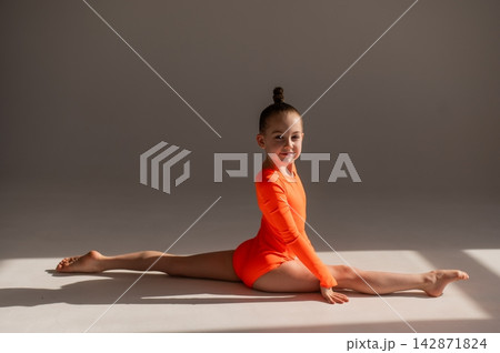Little Caucasian girl gymnast in an orange leotard doing the splits on a white background. Little Caucasian girl gymnast in an orange leotard doing the splits on a white background. 142871824