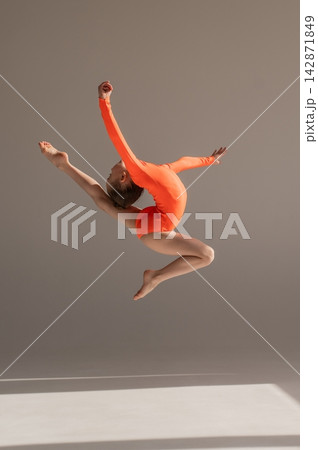 A little Caucasian girl gymnast in an orange leotard demonstrates her skills on a white background.  142871849
