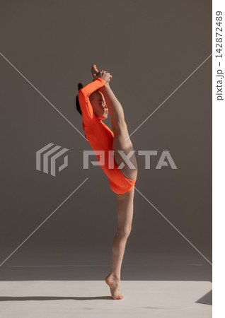 A little Caucasian girl gymnast in an orange leotard demonstrates her skills on a white background. A little Caucasian girl gymnast in an orange leotard demonstrates her skills on a white background. 142872489