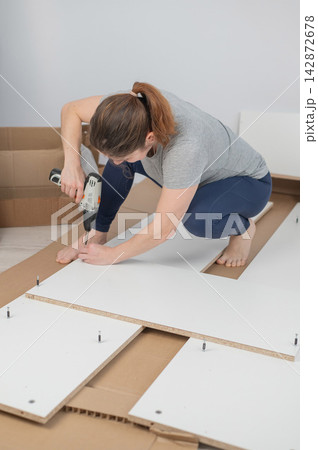 A Caucasian woman uses a screwdriver to tighten a screw while assembling a cabinet. 142872678