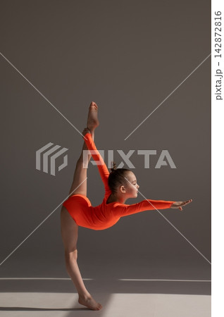 A little Caucasian girl gymnast in an orange leotard demonstrates her skills on a white background. A little Caucasian girl gymnast in an orange leotard demonstrates her skills on a white background. 142872816
