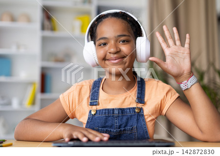 Smiling Black teenage girl wearing headphones waves toward the camera at home Smiling Black teenage girl wearing headphones waves toward the camera at home 142872830