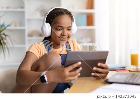 Black teenage girl wearing headphones using tablet at desk at home, Black teenage girl wearing headphones using tablet at desk at home, 142873045