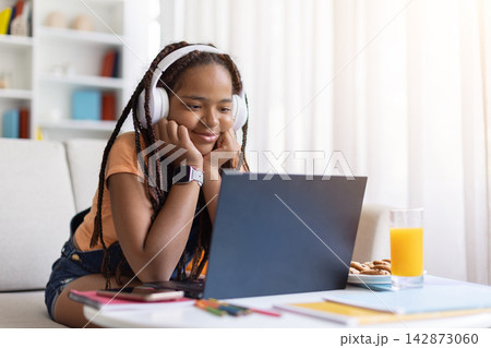 Black teenage girl wearing headphones watching laptop while studying online at home Black teenage girl wearing headphones watching laptop while studying online at home 142873060