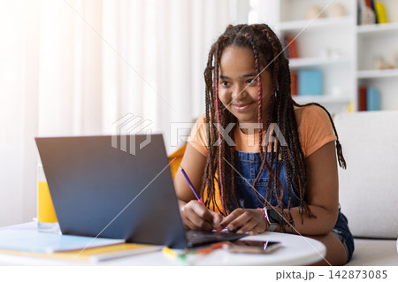 Black teenage girl studying at home with laptop, writing notes with pencil 142873085