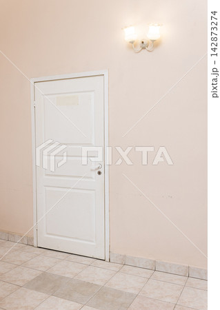 White door against soft pink painted wall with tiled floor and dual bulb wall lamp creating subtle retro interior atmosphere, minimalist design concept and pastel aesthetic background White door against soft pink painted wall with tiled floor and dual bulb wall lamp creating subtle retro interior atmosphere, minimalist design concept and pastel aesthetic background 142873274