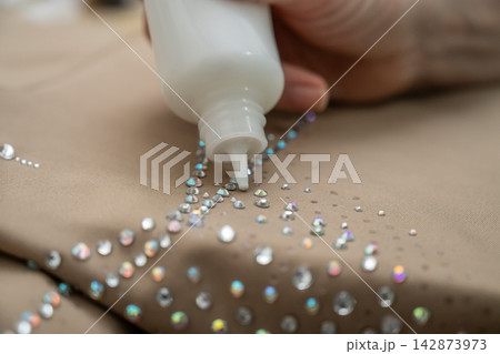 A woman decorates a gymnastics costume with rhinestones. Rhythmic gymnastics. Glue in a tube. A woman decorates a gymnastics costume with rhinestones. Rhythmic gymnastics. Glue in a tube. 142873973