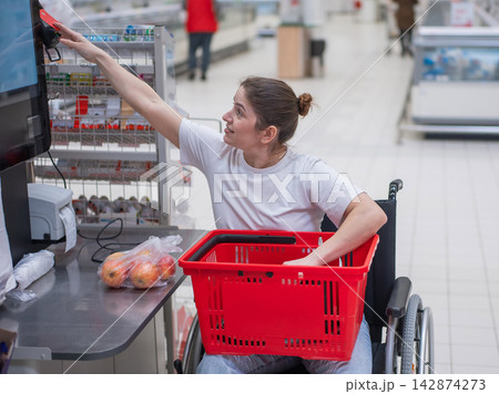 A Caucasian woman in a wheelchair pays for groceries at a self-checkout counter in a supermarket. A Caucasian woman in a wheelchair pays for groceries at a self-checkout counter in a supermarket. 142874273