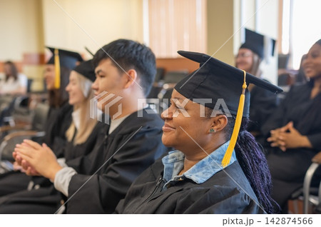 Young people in graduation gowns listen to a speech and applaud. Young people in graduation gowns listen to a speech and applaud. 142874566