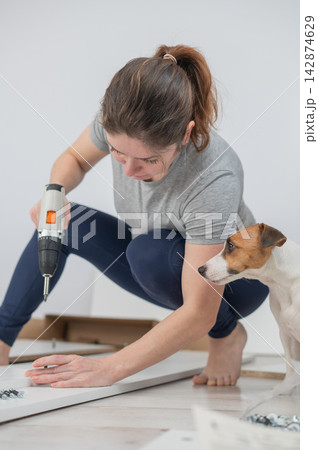 A Caucasian woman uses a screwdriver to tighten a screw while assembling a cabinet. A Caucasian woman uses a screwdriver to tighten a screw while assembling a cabinet. 142874629