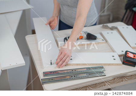 A Caucasian woman uses a screwdriver to tighten a screw while assembling a cabinet. A Caucasian woman uses a screwdriver to tighten a screw while assembling a cabinet. 142874884