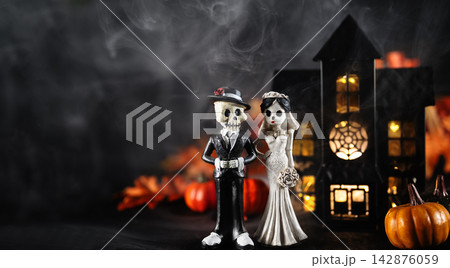 Skeleton bride and groom figurines beside glowing haunted house, pumpkins and smoke. Gothic Halloween wedding decorations on dark background with ample copy space. Halloween holiday card. 142876059