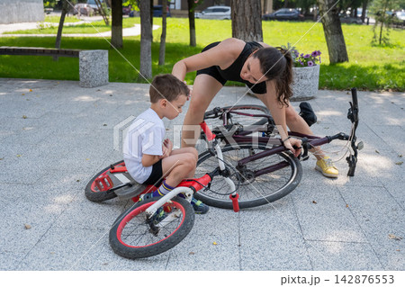 A Caucasian woman is riding a bicycle with her son. The boy fell, and the mother feels sorry for him. A Caucasian woman is riding a bicycle with her son. The boy fell, and the mother feels sorry for him. 142876553