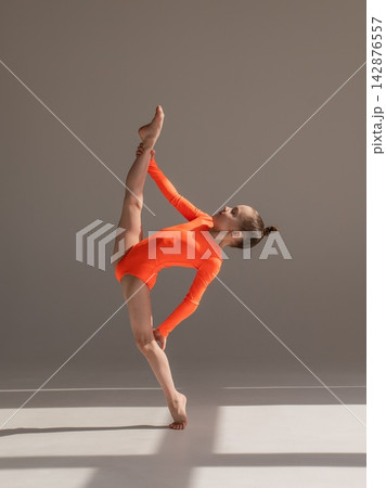 A little Caucasian girl gymnast in an orange leotard demonstrates her skills on a white background. A little Caucasian girl gymnast in an orange leotard demonstrates her skills on a white background. 142876557