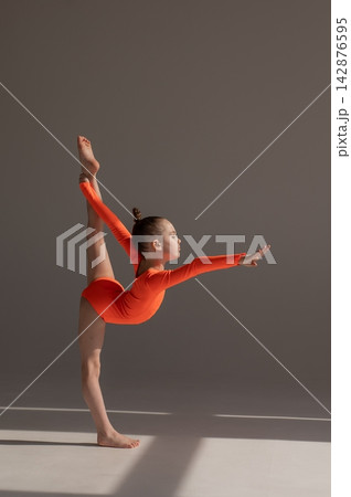A little Caucasian girl gymnast in an orange leotard demonstrates her skills on a white background. A little Caucasian girl gymnast in an orange leotard demonstrates her skills on a white background. 142876595