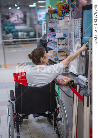 A Caucasian woman in a wheelchair pays for groceries at a self-checkout counter in a supermarket. A Caucasian woman in a wheelchair pays for groceries at a self-checkout counter in a supermarket. 142876648