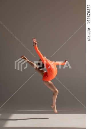 A little Caucasian girl gymnast in an orange leotard demonstrates her skills on a white background. A little Caucasian girl gymnast in an orange leotard demonstrates her skills on a white background. 142876872