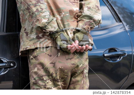 Close view of detained man in camouflage with hands secured behind his back in metal handcuffs beside a vehicle. Close view of detained man in camouflage with hands secured behind his back in metal handcuffs beside a vehicle. 142878354