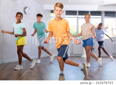 Positive juvenile boy engaged in Rock 'n' Roll dance in training room with children's group Positive juvenile boy engaged in Rock 'n' Roll dance in training room with children's group 142879743