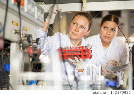 Female researchers in lab coats writing notes and looking at reaction in test tubes during biochemical tests in laboratory 142879950