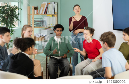 Teen pupils gathering in circle, participate at speaking club with coach, teacher watching. 142880001