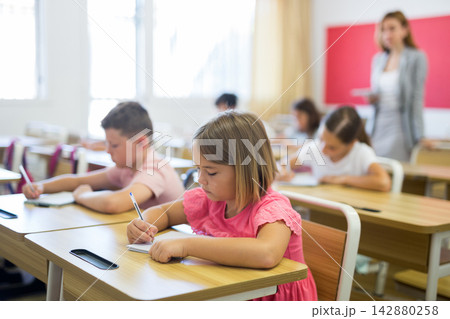 Interested blonde tween girl listening to schoolteacher and writing in notebook Interested blonde tween girl listening to schoolteacher and writing in notebook 142880258