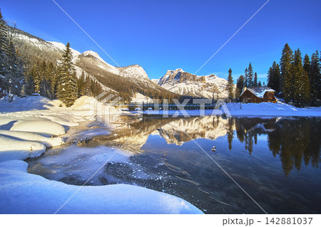 Winter Lodge Reflections and Frozen Lake with Snowy Mountains under Clear Blue Sky Winter Lodge Reflections and Frozen Lake with Snowy Mountains under Clear Blue Sky 142881037