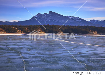 Frozen Lake with Cracked Ice Pines and Rugged Mountain Peaks in Winter 142881308