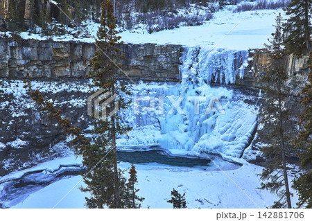 Frozen Waterfall and Icy Pool with Cliff Face and Evergreens in Winter Landscape 142881706