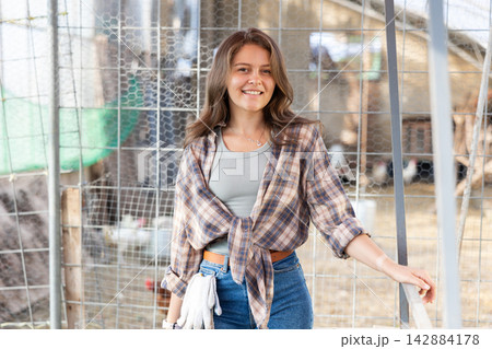 Young woman owner of livestock farm stands near corral mesh fence for livestock and rests Young woman owner of livestock farm stands near corral mesh fence for livestock and rests 142884178
