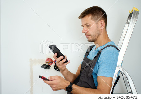 Man using smartphone during wall outlet installation 142885472
