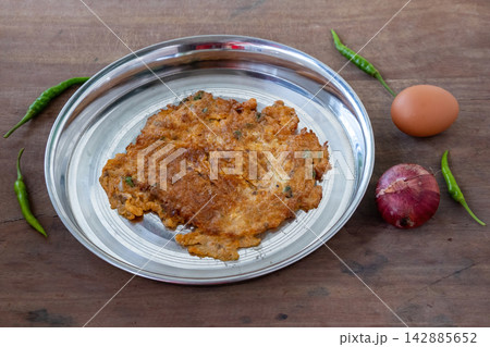 Crispy Fried Egg Omelet Cooked with Onions and Green Chilies, Served on Stainless Steel Plate with Whole Egg, Onion, Green Chili Props, Wooden Background. Crispy Fried Egg Omelet Cooked with Onions and Green Chilies, Served on Stainless Steel Plate with Whole Egg, Onion, Green Chili Props, Wooden Background. 142885652