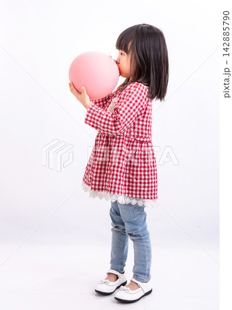 Cute little Asian girl in a red plaid shirt blowing up a pink balloon, standing against a white background, childhood fun and party preparation concept. Cute little Asian girl in a red plaid shirt blowing up a pink balloon, standing against a white background, childhood fun and party preparation concept. 142885790