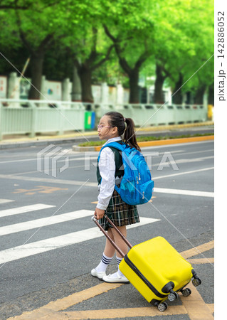 Asian schoolgirl in a uniform carrying a blue backpack and holding a yellow suitcase, standing safely at a pedestrian zebra crossing, school road safety education concept. Asian schoolgirl in a uniform carrying a blue backpack and holding a yellow suitcase, standing safely at a pedestrian zebra crossing, school road safety education concept. 142886052